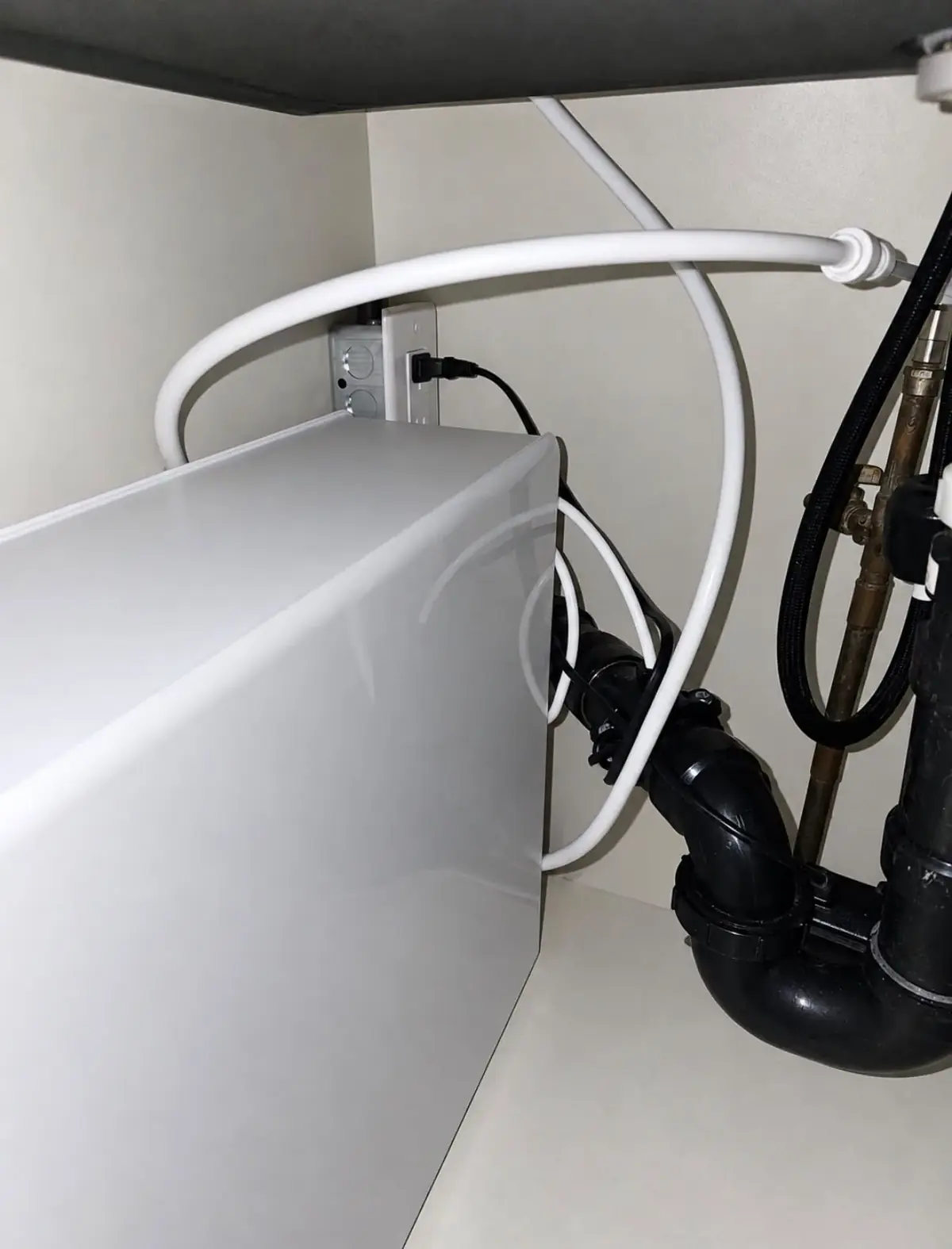 Receptacle mounted on the back wall of a sink cabinet with a cord plugged in, beside a water treatment unit, white tubing and a black drain trap