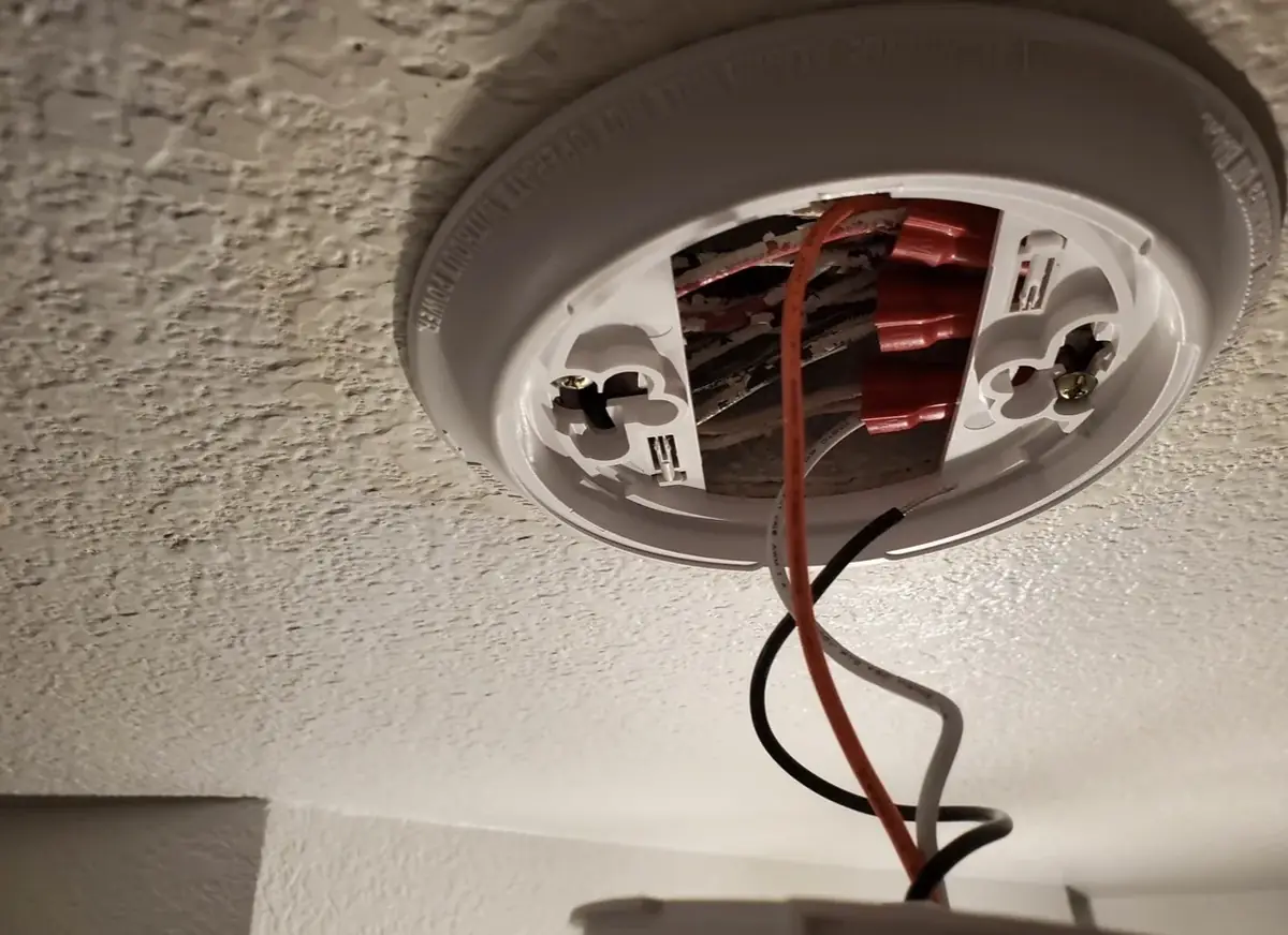 Smoke alarm mounting base open on a textured ceiling, with red, black and white conductors joined by wire connectors