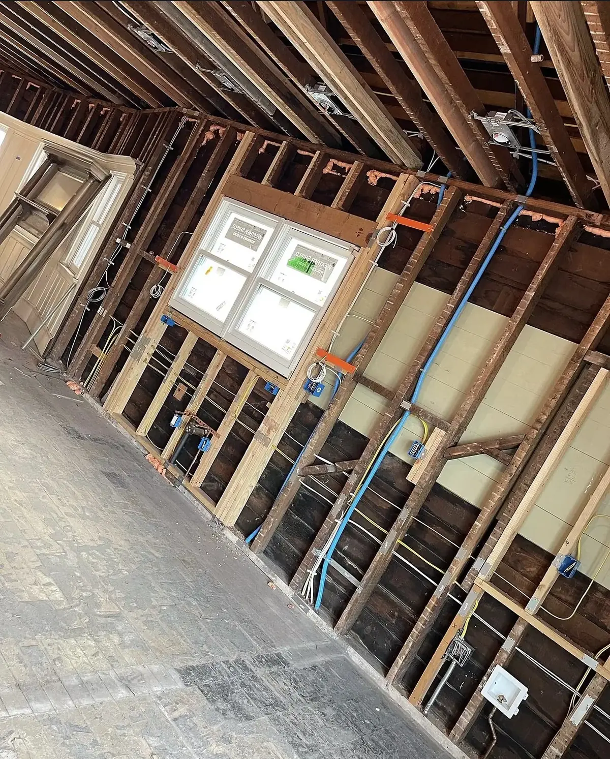 Gutted room with exposed wood studs and two windows, yellow and blue cables run to blue device boxes at receptacle height and recessed light housings between the ceiling joists