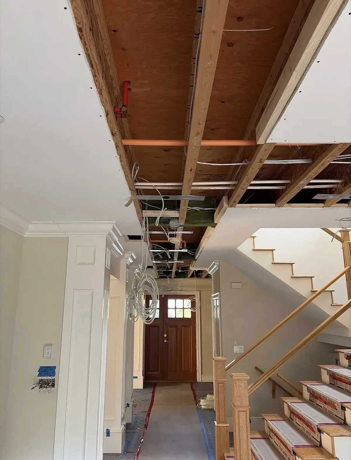 Hallway with the ceiling drywall removed exposing joists, cables and plumbing, a staircase on the right and the front door at the end