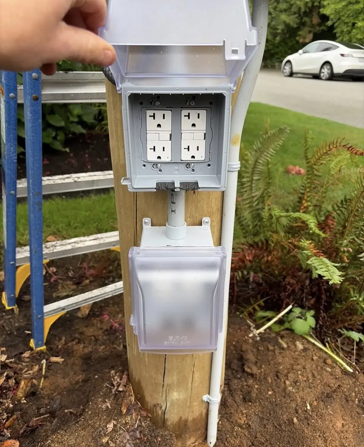 Weatherproof in-use cover held open to show two receptacles mounted on a wooden post with PVC conduit, a second covered outlet below and a garden bed behind