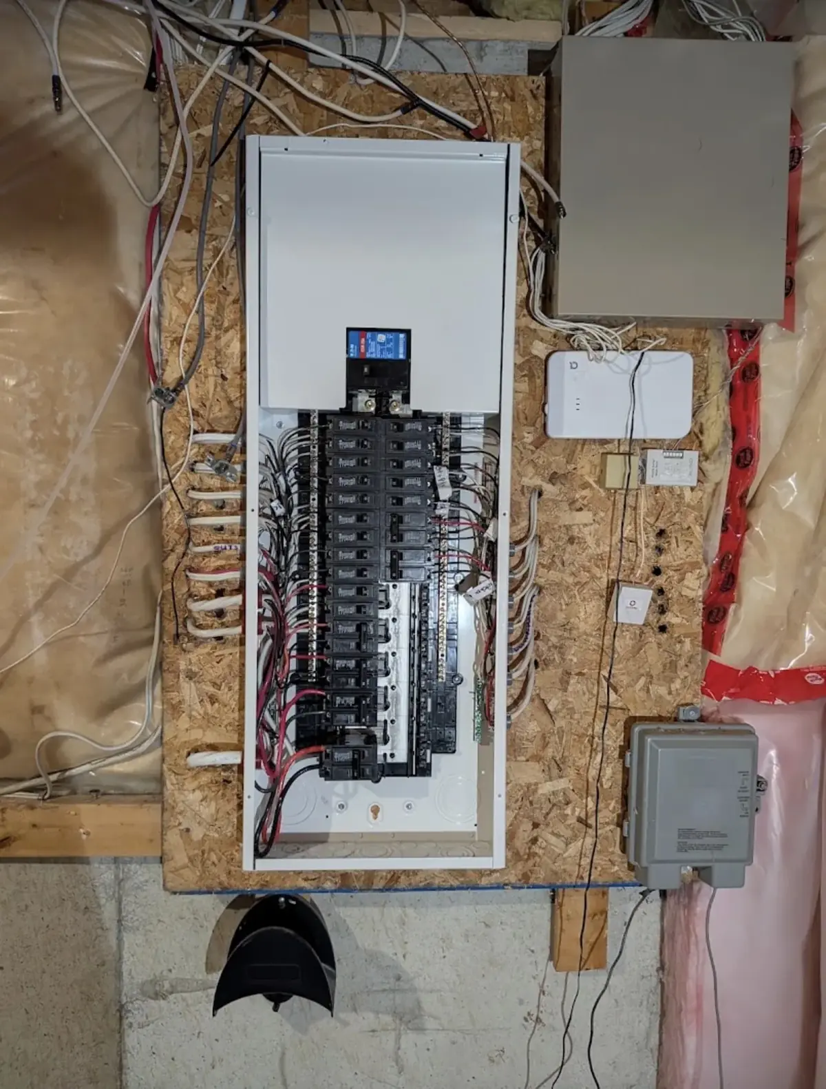 Breaker panel with the cover removed, mounted on a plywood backboard in an unfinished basement, white conductors dressed along the sides and surface-mounted boxes above and below
