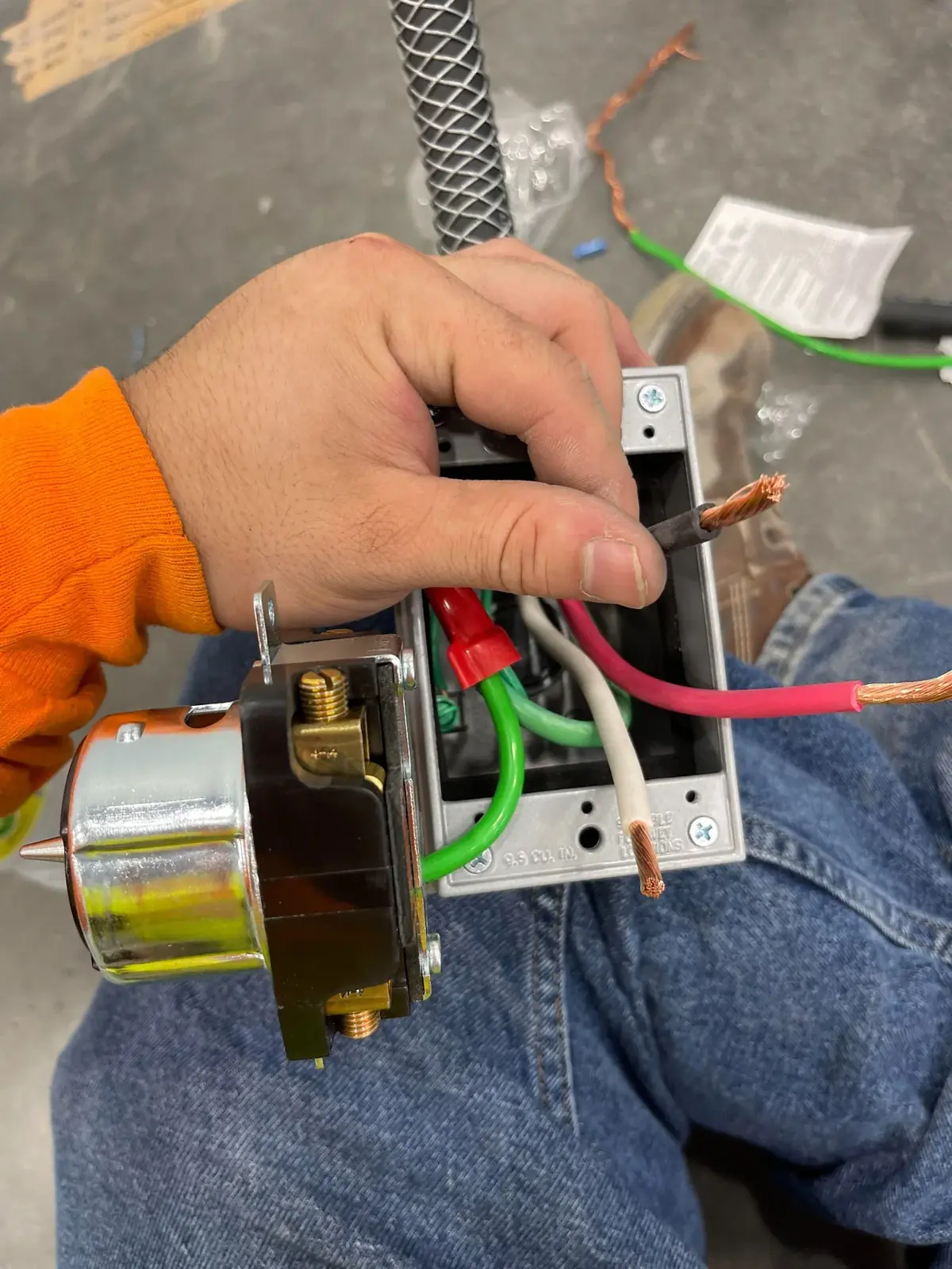 Hand holding a four-wire receptacle with its terminals exposed in front of a metal device box, with red, black, white and green conductors stripped for termination