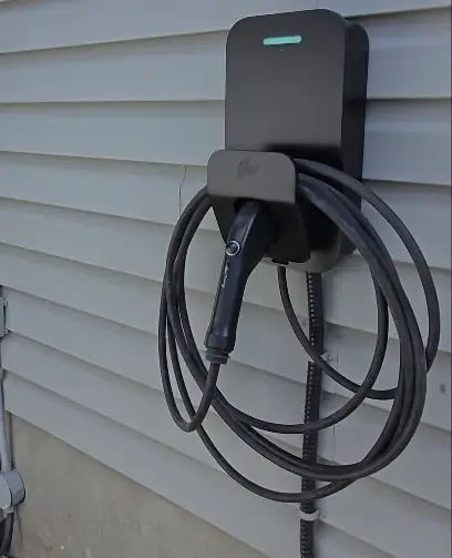 Black EV charger mounted on grey exterior siding with the cable coiled on its holster