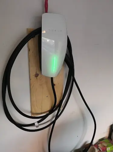 White EV wall connector mounted on a wood board in a garage with the cable coiled around it