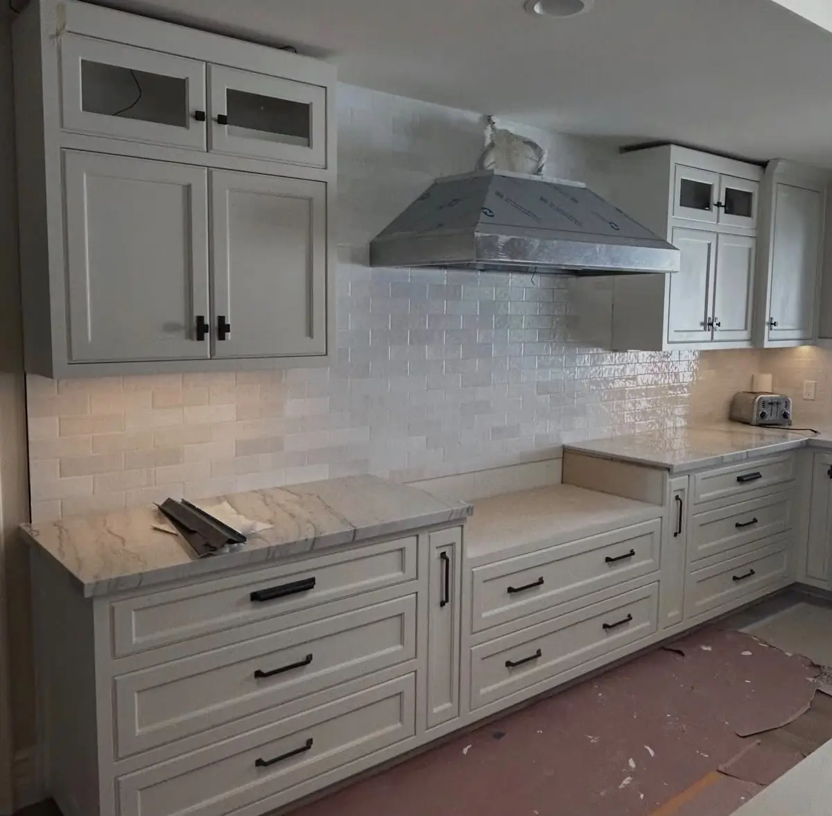 White kitchen cabinets with under-cabinet lighting on, a wrapped range hood and construction dust on the floor